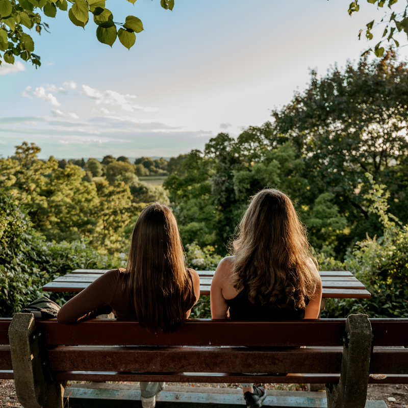 Twee jonge vrouwen met hun rug naar ons toe op een bankje uitkijkend over groene omgeving