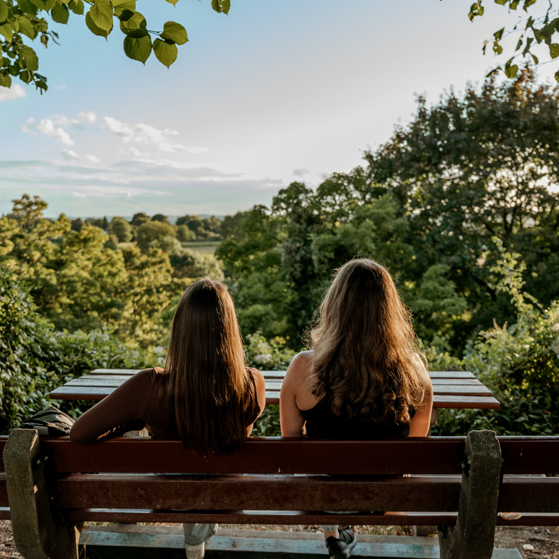 Twee jonge vrouwen met hun rug naar ons toe op een bankje uitkijkend over groene omgeving